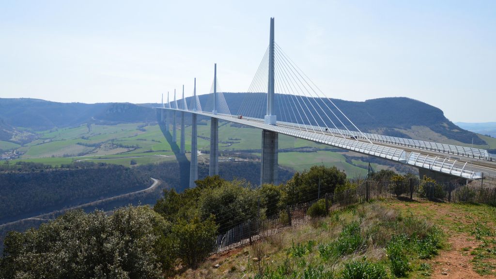 Viaduto estaiado de pista dupla atravessando um vale largo, com mastros altos e cabos em leque, campos verdes e montanhas ao fundo