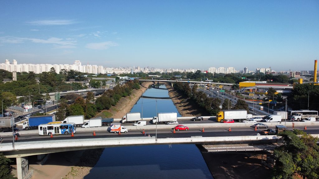 Caminhões, ônibus e carros numa via expressa que cruza um rio canalizado, com a cidade ao fundo