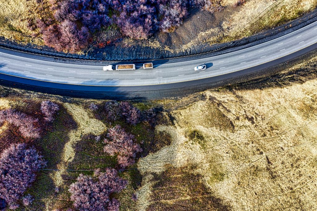 Rodovia em curva vista do alto, entre mato seco, com um caminhão de dois reboques e um carro em movimento