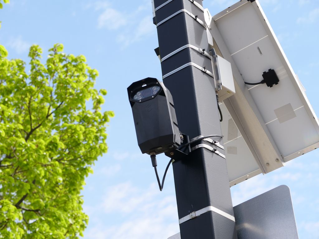Câmera de leitura de placa com iluminador presa a um poste, ao lado de um painel solar, contra o céu azul