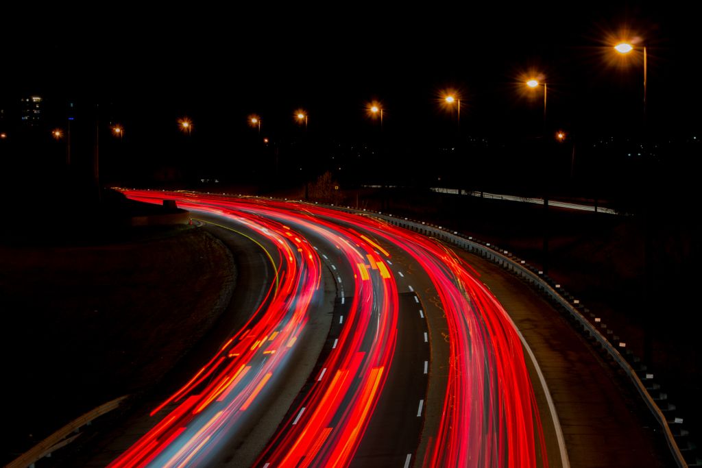 Rodovia em curva à noite, com os rastros de luz de faróis e lanternas