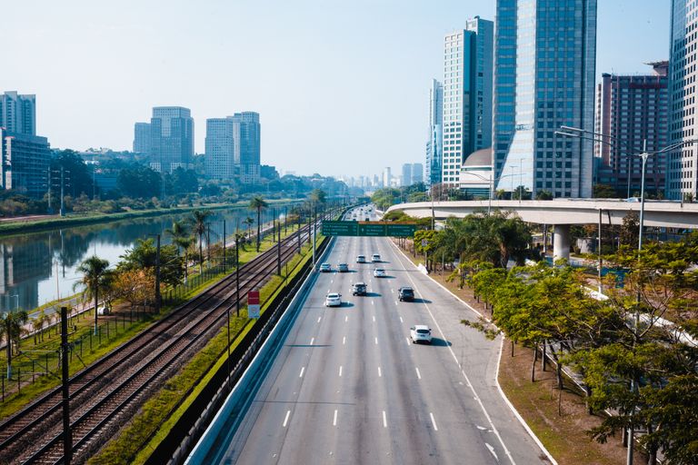 Via expressa urbana de várias faixas ao lado de uma linha de trem e de um rio, com viaduto e prédios ao fundo
