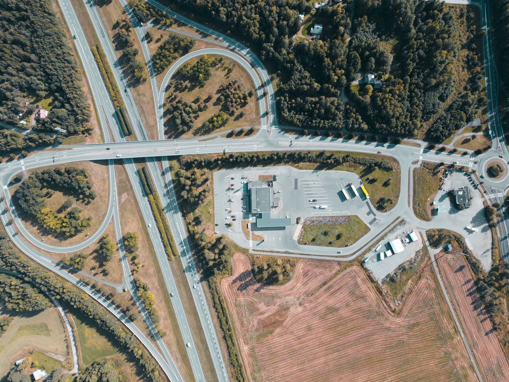 Trevo rodoviário visto de cima, com um empreendimento e seu estacionamento ao lado da rodovia e a via local que segue até uma rotatória