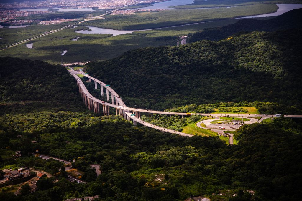 Viadutos de pista dupla em curva sobre a mata da serra, com um trevo ao lado, vistos do alto