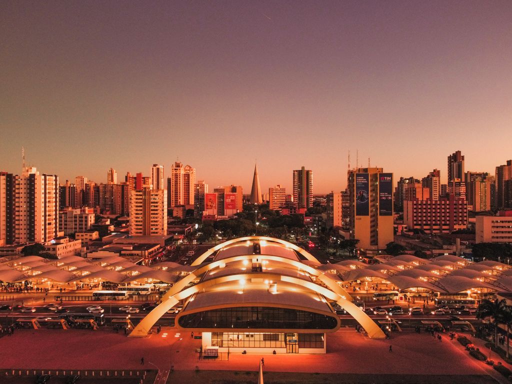 Terminal de ônibus ao entardecer, com as coberturas em arco acesas e os prédios ao fundo